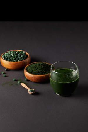 wooden spoon, glass of fresh spirulina smoothie, bowls with spirulina powder and spirulina pills on grey tableの写真素材