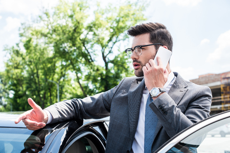 businessman in eyeglasses talking on smartphone while standing near car on streetの写真素材