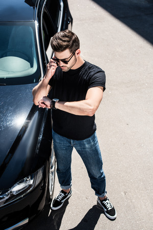 high angle view of stylish young man in sunglasses checking wristwatch near his car at streetの写真素材