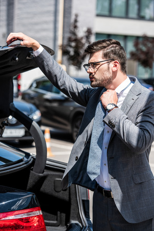 young businessman in eyeglasses closing car trunk at streetの写真素材