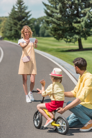 happy young woman with smartphone photographing father teaching daughter riding bicycle in parkの写真素材