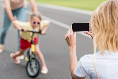 woman with smartphone photographing father and daughter riding bicycle in parkの写真素材