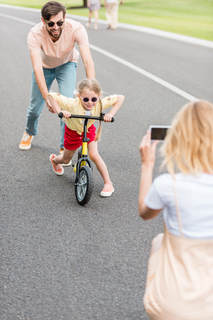 woman with smartphone photographing happy father and daughter riding bicycle in parkの写真素材