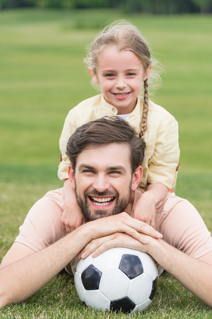 happy father and daughter smiling at camera while lying with soccer ball on grassの写真素材