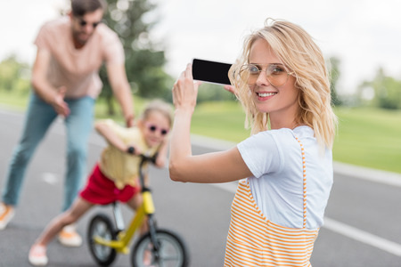 woman holding smartphone and smiling at camera while photographing father teaching daughter riding bicycleの写真素材