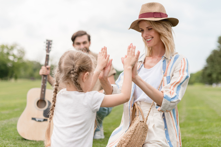 happy mother and daughter playing together while father holding acoustic guitar behind in parkの写真素材