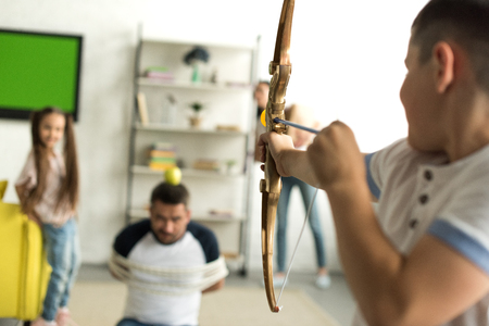 siblings playing with tied father and pretending shooting with toy bow at homeの写真素材