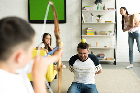 children playing with tied father and pretending shooting with toy bow in living room