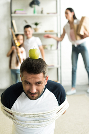 tied father sitting on floor with apple on head for target at home, parenthood conceptの写真素材