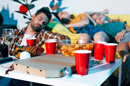 close-up view of pizza box, plastic cups and baseball equipment on table and drunk young people sleeping behindの写真素材