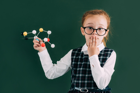 shocked little schoolchild holding molecular model and looking at camera while standing near chalkboardの写真素材