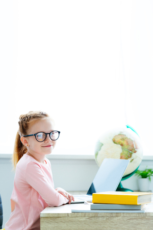 beautiful red haired schoolgirl in eyeglasses smiling at camera while studying with books and laptop at deskの写真素材