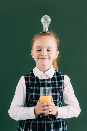 happy little schoolgirl with closed eyes and light bulb on head holding glass of orange juiceの写真素材