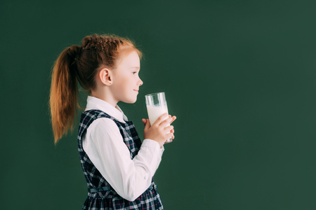 side view of smiling little schoolgirl holding glass of milk while standing near chalkboardの写真素材