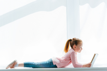 side view of cute little schoolgirl lying on windowsill and reading bookの写真素材