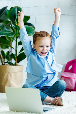 cheerful little schoolgirl raising hands while studying with laptop at homeの写真素材