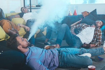 young man smoking electronic cigarette while drunk friends sleeping after party