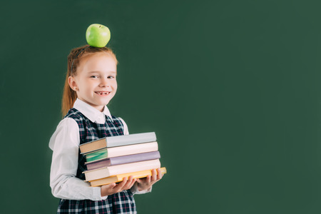 beautiful little schoolgirl with apple on head holding pile of books and smiling at cameraの写真素材