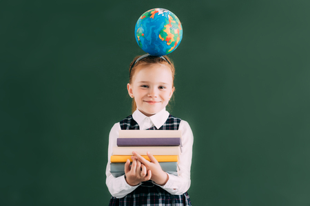 little redhead schoolgirl with globe on head holding pile of books and smiling at cameraの写真素材