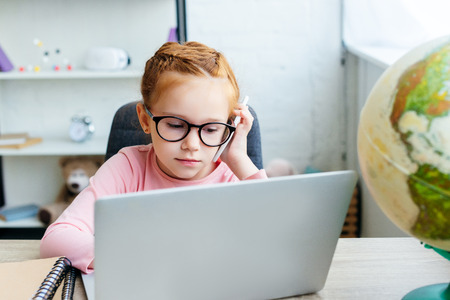 focused child in eyeglasses talking by smartphone and using laptop at home