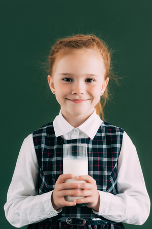 adorable little schoolgirl holding glass of milk and smiling at cameraの写真素材