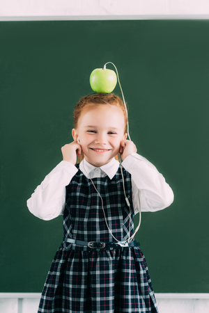 adorable little schoolgirl with apple on head and earphones smiling and looking at cameraの写真素材