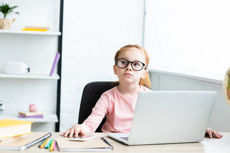thoughtful little schoolchild in eyeglasses looking up while sitting at desk and using laptop