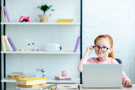 adorable red haired schoolgirl in eyeglasses using laptop and smiling at camera while studying at homeの写真素材