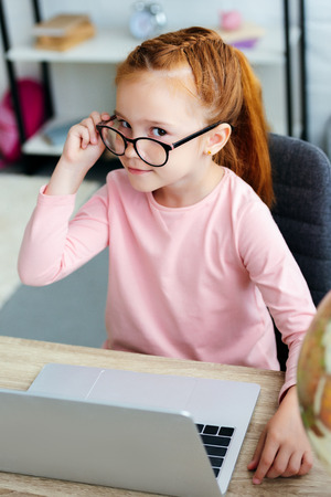 high angle view of beautiful schoolgirl adjusting eyeglasses and smiling at camera while using laptopの写真素材