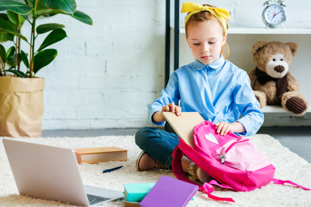 cute little child packing school bag while sitting on carpet at home