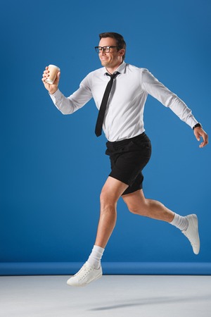 happy man holding disposable coffee cup while jumping and looking away on blueの写真素材
