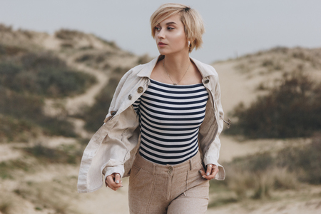 beautiful young woman in stylish striped shirt and vintage jacket walking on sand dunesの写真素材