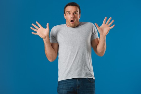 shocked man raising hands and looking at camera isolated on blue