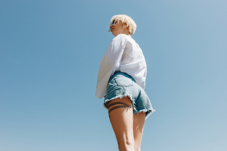 low angle view of beautiful young stylish woman in sunglasses and denim shorts standing against clear blue skyの写真素材