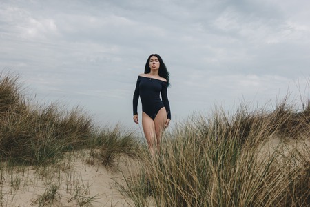 attractive young woman in black bodysuit on sand dune under cloudy skyの写真素材