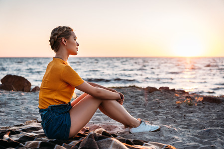 side view of pensive young woman sitting on plaid at beautiful sea coast at sunsetの写真素材