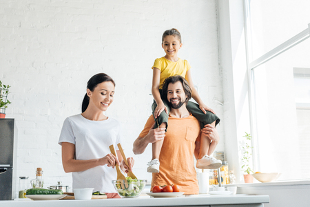 smiling young woman preparing salad while her daughter riding on shoulders of husband at kitchenの写真素材