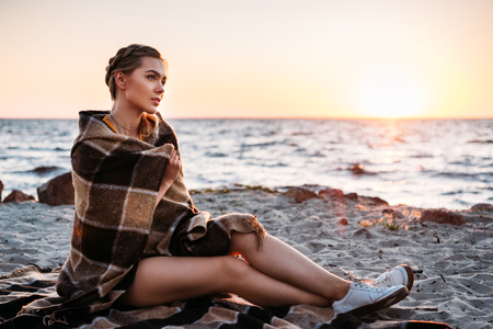 beautiful pensive young woman sitting wrapped in plaid and looking at majestic sunset at seaの写真素材