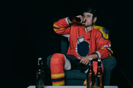 young hockey player drinking beer while sitting in armchair and watching tv on black