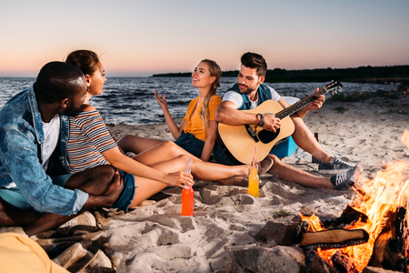 happy young multiethnic friends enjoying guitar and spending time together on sandy beach at sunsetの写真素材
