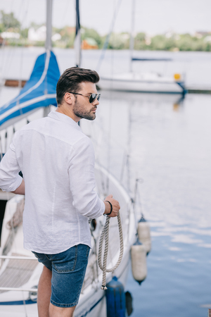 handsome young man in sunglasses looking away while holding rope at yachtの写真素材