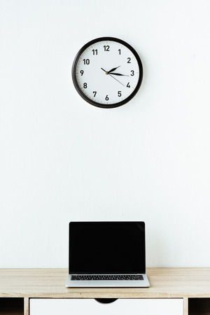 laptop on work desk under clock hanging on white wallの写真素材