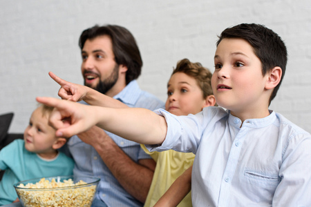 excited man and little sons with popcorn pointing away while watching film together at home