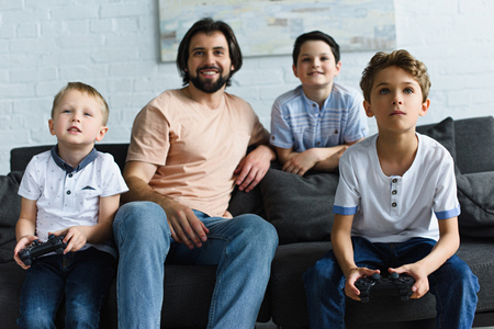 smiling father looking at little sons sitting on sofa and playing video games together at homeの写真素材