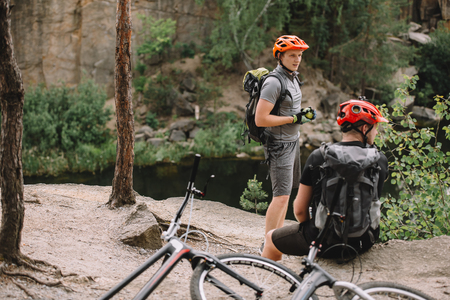 two male friends with backpacks and bicycles resting near river in forestの写真素材