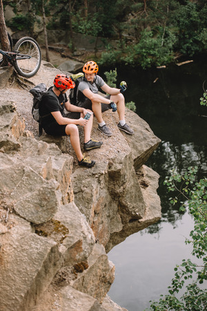 high angle view of friends cyclists with backpacks resting with sport bottle of water and apple on rocky cliff near river in forestの写真素材