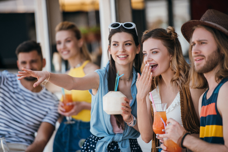 happy young friends holding beverages and looking away while spending time together at beach barの写真素材