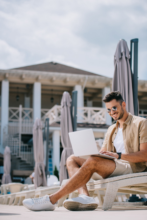 smiling young man in sunglasses sitting on chaise longue and using laptopの写真素材