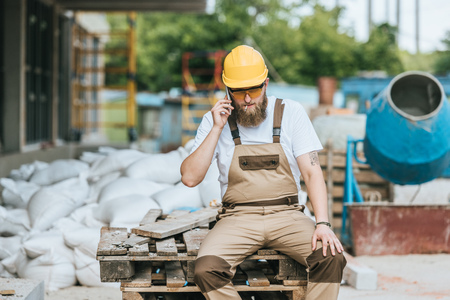 builder in hardhat and protective goggles smoking cigarette and talking on smartphone at construction siteの写真素材
