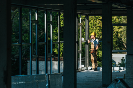 distant view of builder in protective helmet and goggles talking on smartphone at construction site